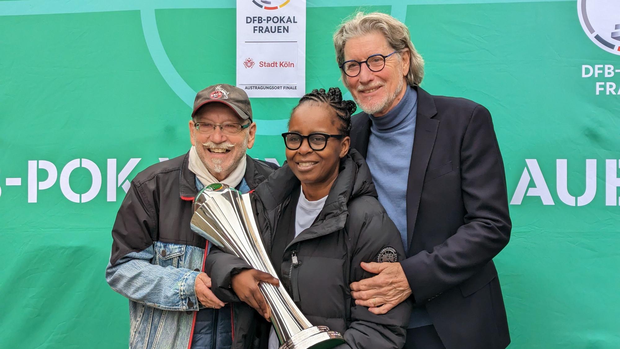 Janus Fröhlich, Shary Reeves und Toni Schumacher stehen zusammen vor einem grünen Hintergrund. Shary Reeves hat den Pokal in der Hand.