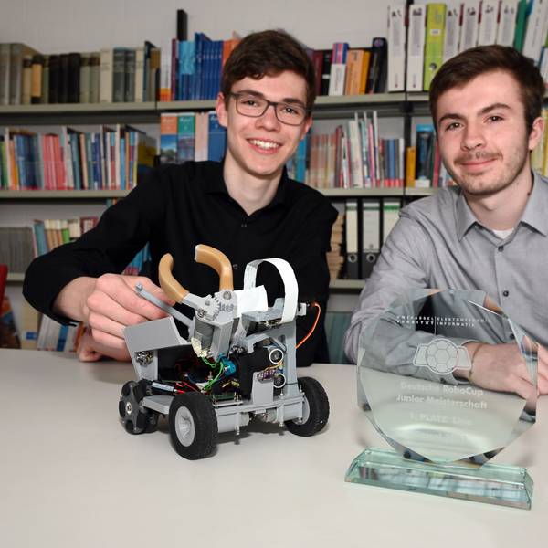 Zwei Schüler sitzen an einem Tisch, vor ihnen stehen ein kleiner Roboter und eine Siegertrophäe für einen Roboter-Wettbewerb auf dem Tisch.