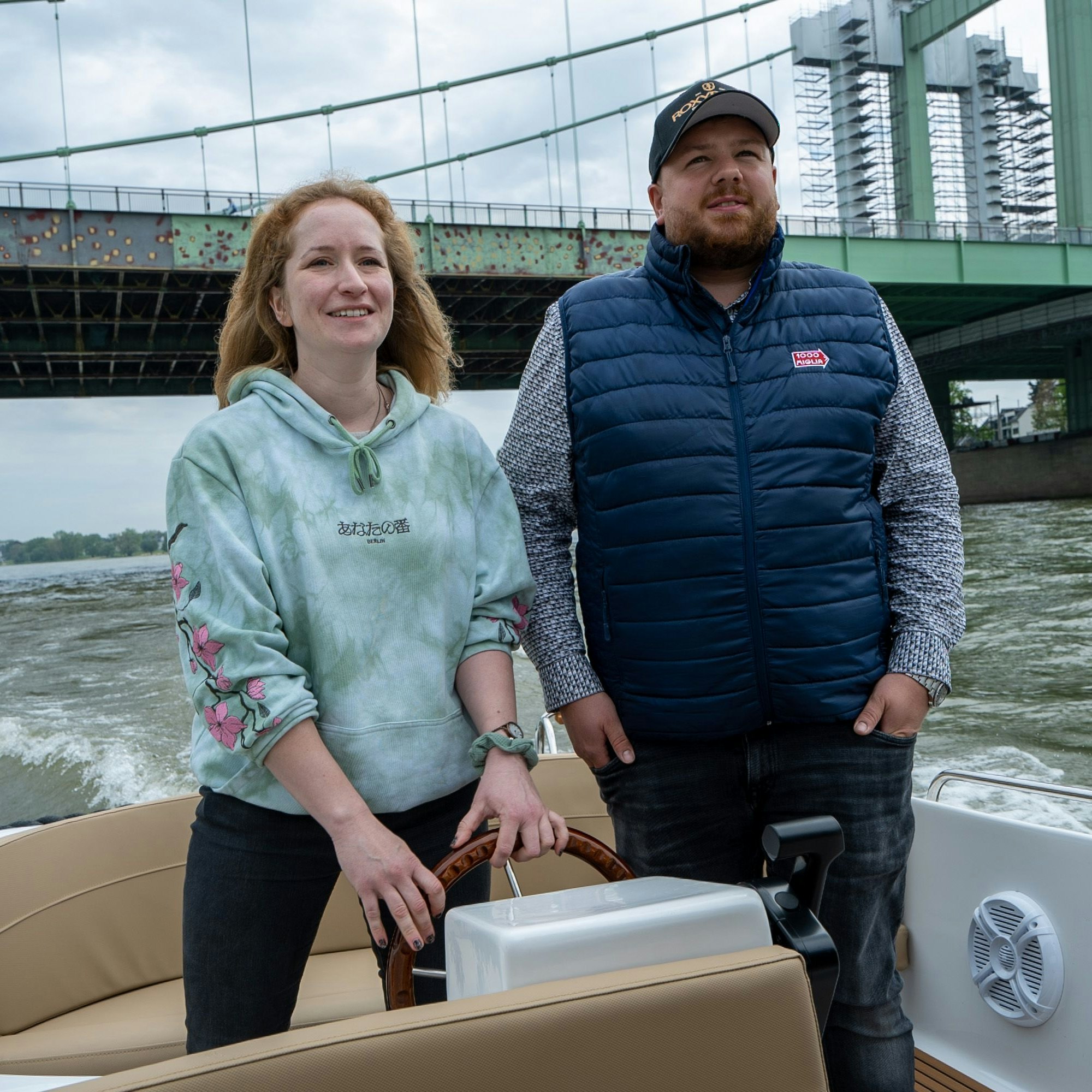 Eine Frau und ein Mann stehen am Steuer des Bootes auf dem Rhein, im Hintergrund die Severinsbrücke.