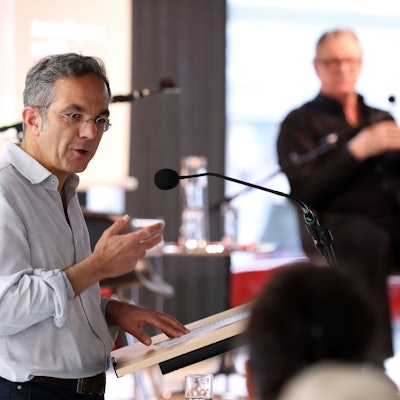 Kermani hat ein Hemd an, das er am Ärmel leicht hochgekrämeptk hat. Er steht an einem Punkt und redet in ein Mikrofon, im Hintergrund eine verschwommene Person.