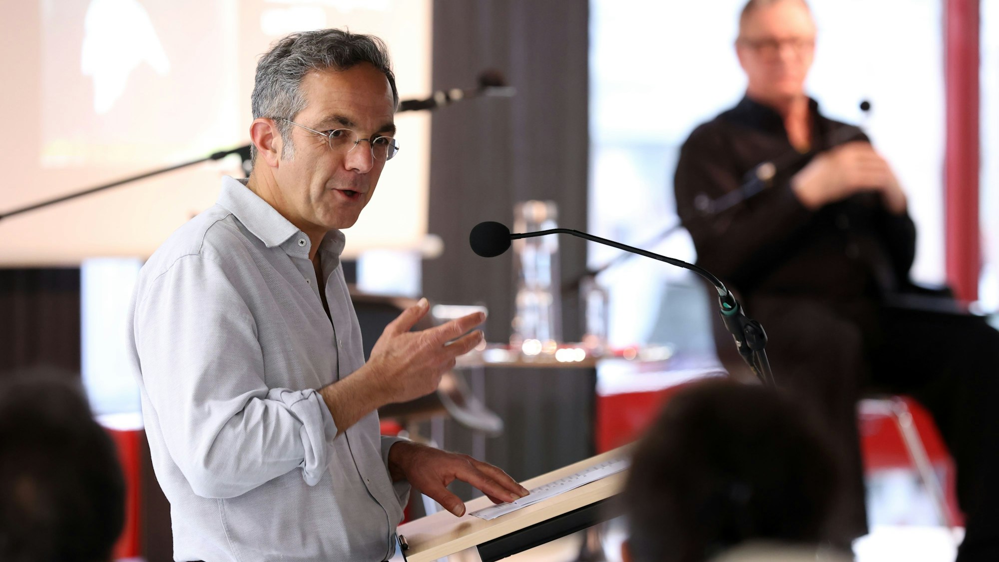 Kermani hat ein Hemd an, das er am Ärmel leicht hochgekrämeptk hat. Er steht an einem Punkt und redet in ein Mikrofon, im Hintergrund eine verschwommene Person.