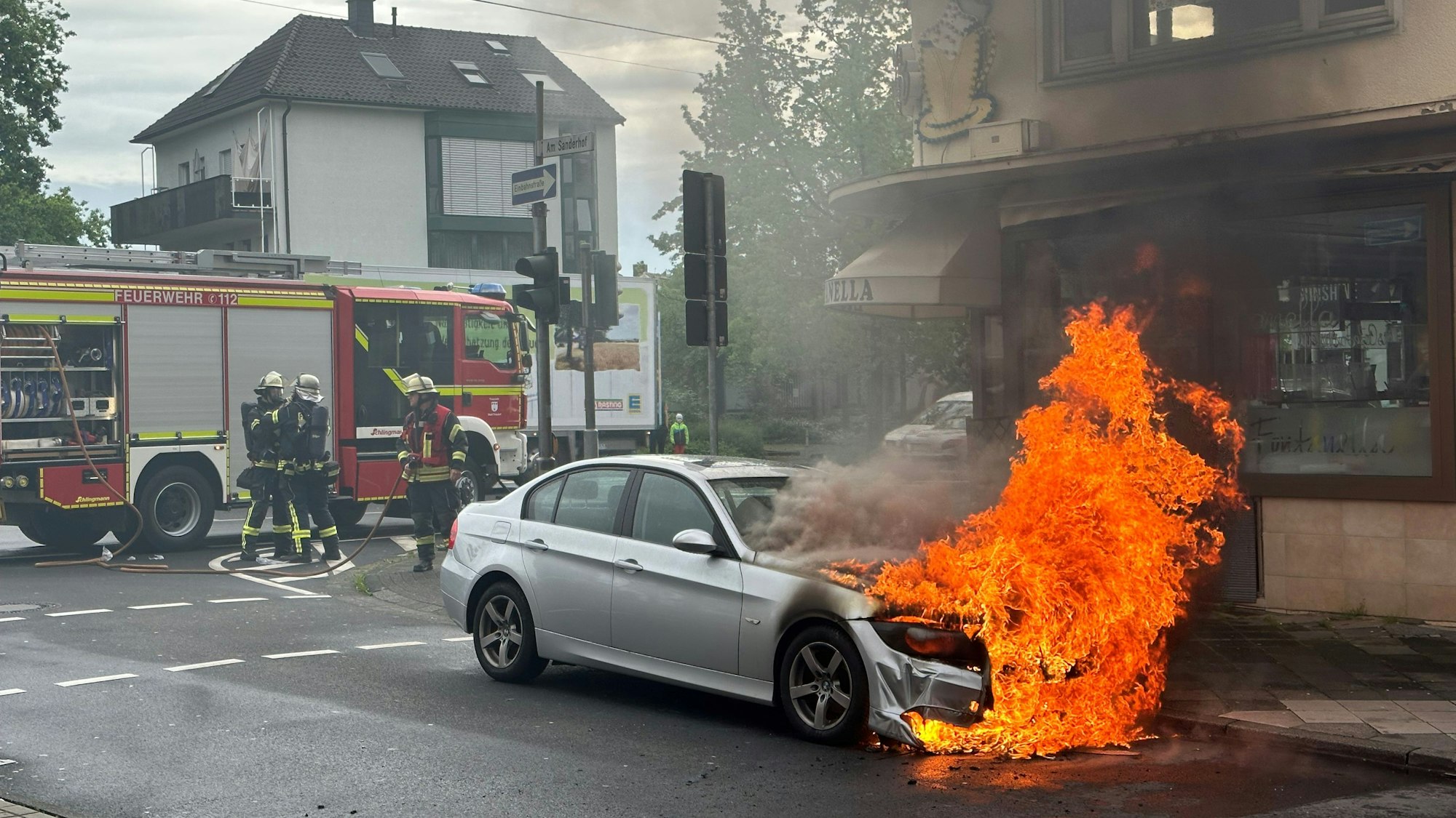 Der Motorraum eines silbernen Autos steht lichterloh in Flammen. Schwarzer Rauch zieht auf. Im Hintergrund bereiten Feuerwehrleute die Löscharbeiten vor.