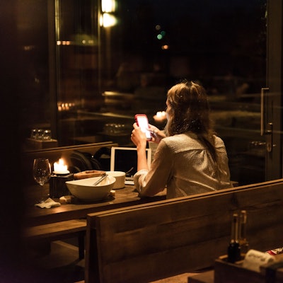 Eine Frau sitzt in einem relativ dunklen und weitestgehend leeren Restaurant an einem Tisch mit leerem Teller und schaut auf ihr Smartphone.