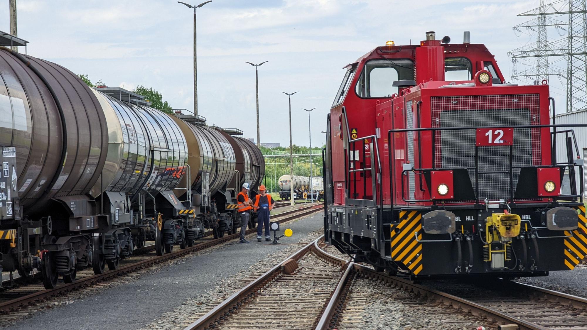 Der Chemiepark Knapsack in Hürth. Zwischen Gleisen und zwei Zügen stehen zwei Gleisarbeiter in orangenen Westen mit Bauhelmen.