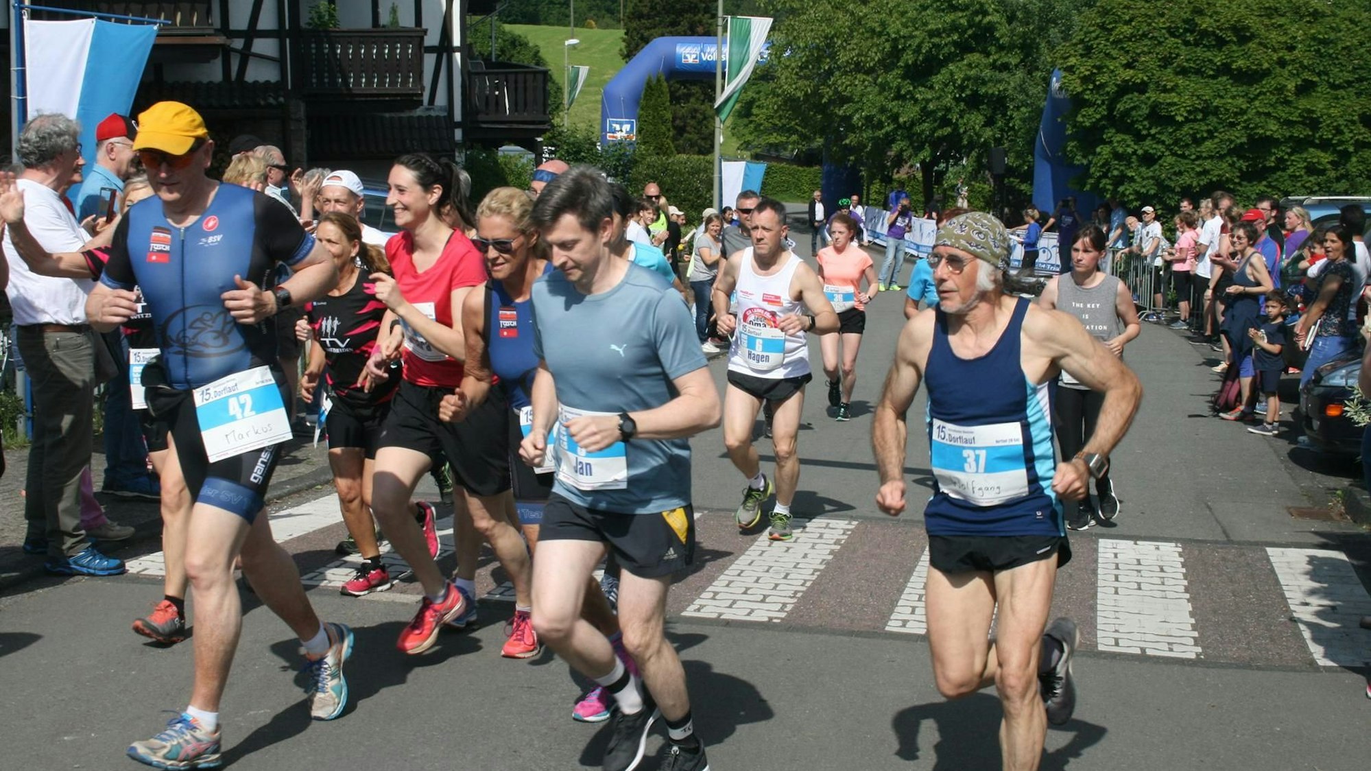 Der Biesfelder Dorflauf 2019. Mehrere Läufer und Läuferinnen in Sportklamotten sind zu sehen. Publikum feuert die Athleten und Athletinnen an.