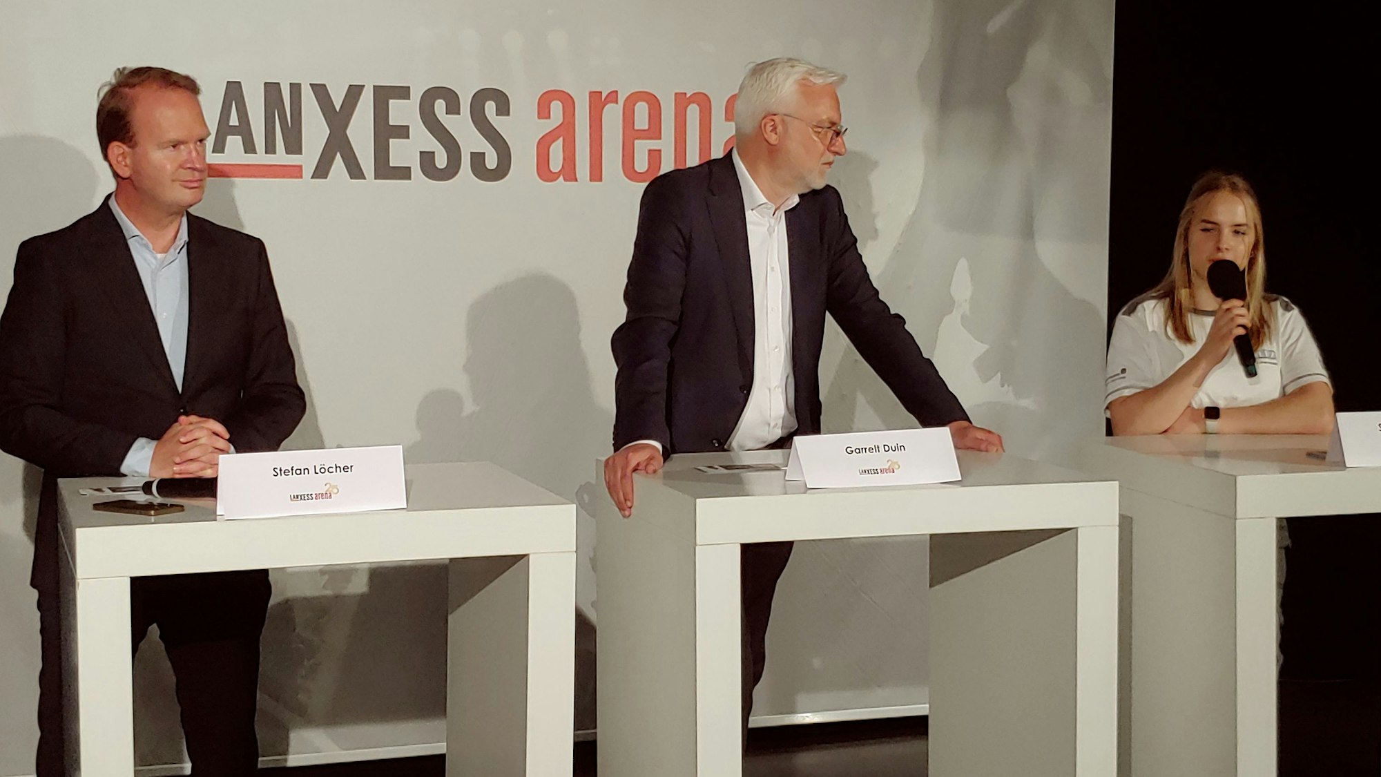 Die drei stehen an weißen Pulten auf einer Bühne