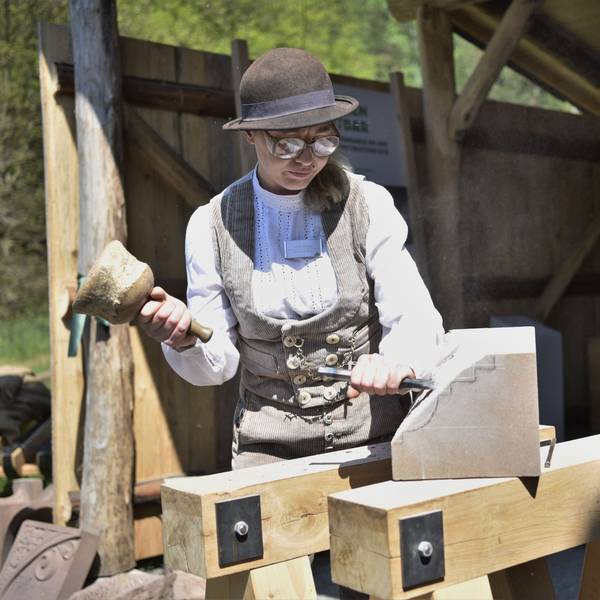 Steinmetzin Ulrike Glaubitz steht vor der Steinmetzhütte im Freilichtmuseum Kommern. Sie trägt traditionelle, beige Handwerks-Kluft und bearbeitet einen Stein mit Hammer und Meißel.