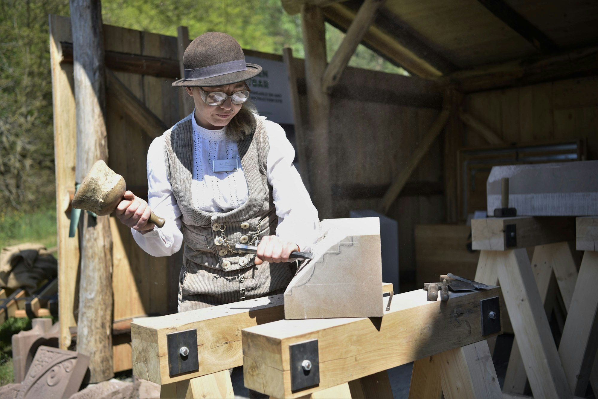 Steinmetzin Ulrike Glaubitz steht vor der Steinmetzhütte im Freilichtmuseum Kommern. Sie trägt traditionelle, beige Handwerks-Kluft und bearbeitet einen Stein mit Hammer und Meißel.