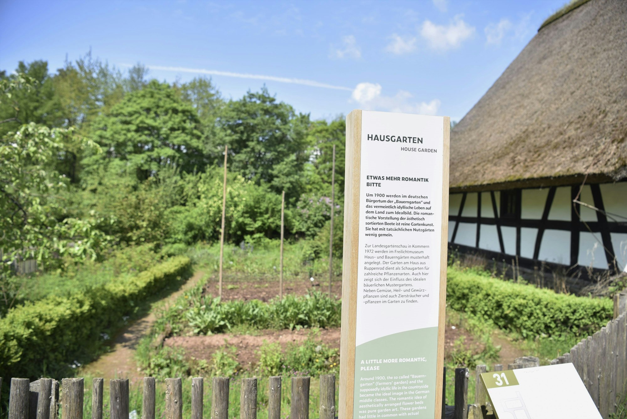 Im Freilichtmuseum Kommern steht eine Informationsstele vor einem angelegten Garten. Die Stele informiert über den Hausgarten.