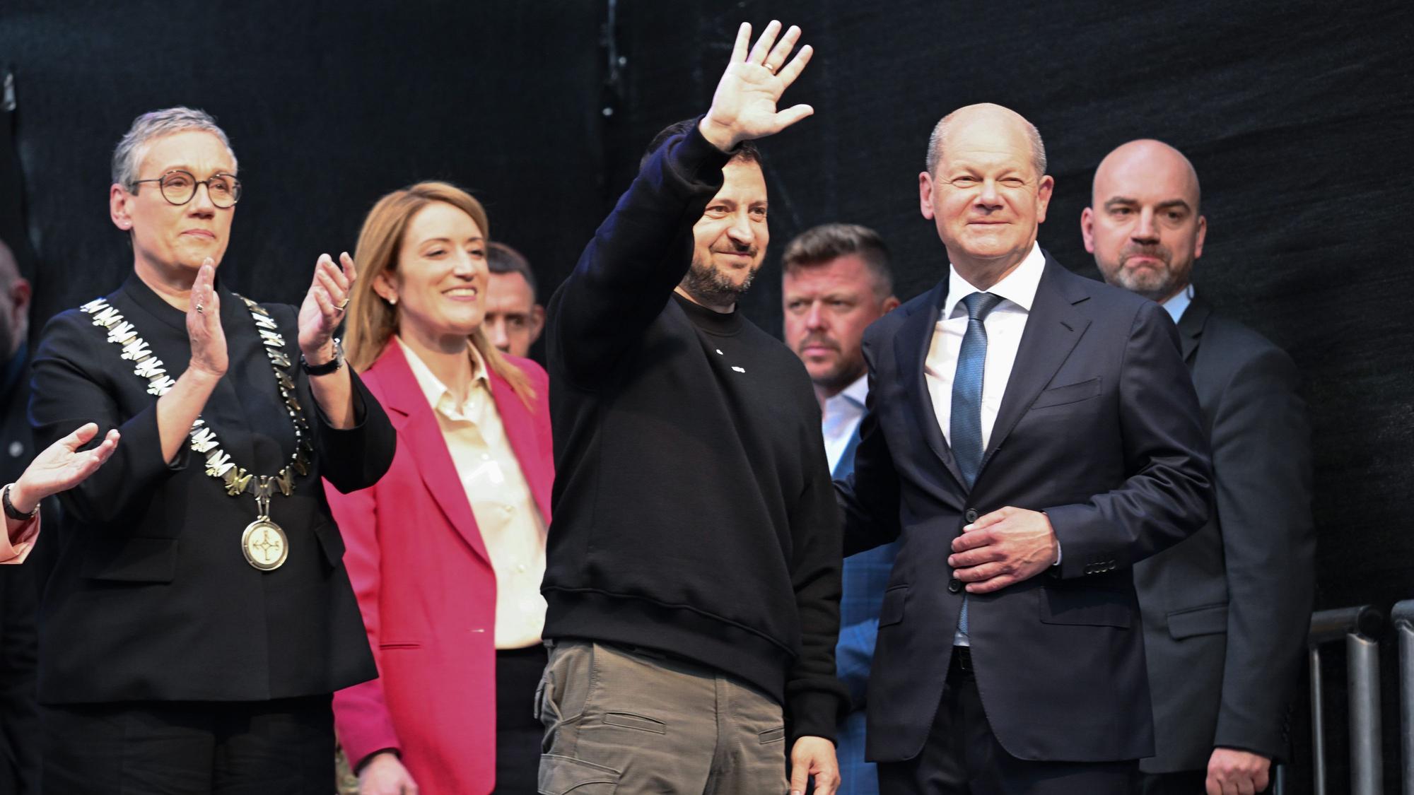 Der ukrainische Präsident und Preisträger Wolodymyr Selenskyj (M) bekommt Applaus und winkt den Menschen vor dem Rathaus nach der Verleihung des Karlspreises für Verdienste um die Einheit Europas. Links Oberbürgermeisterin Sibylle Keupen (parteilos) und Roberta Metsola, EU-Parlamentspräsidentin, 2.v.r Bundeskanzler Olaf Scholz (SPD).