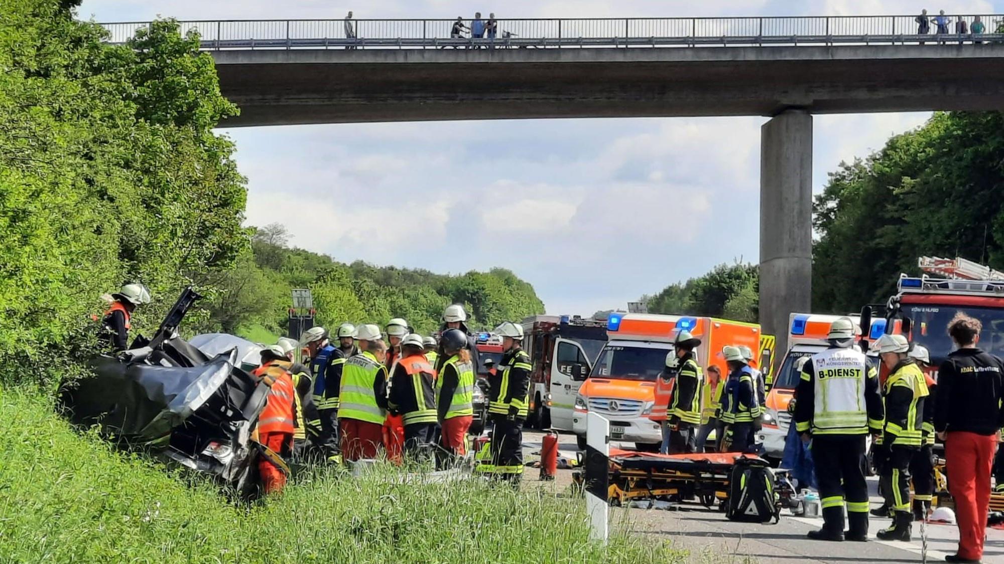 Dicht gedrängt stehen Rettungskräfte um ein Autowrack, das in der Böschung neben der Autobahn liegt. Auf einer Brücke stehen Schaulustige.