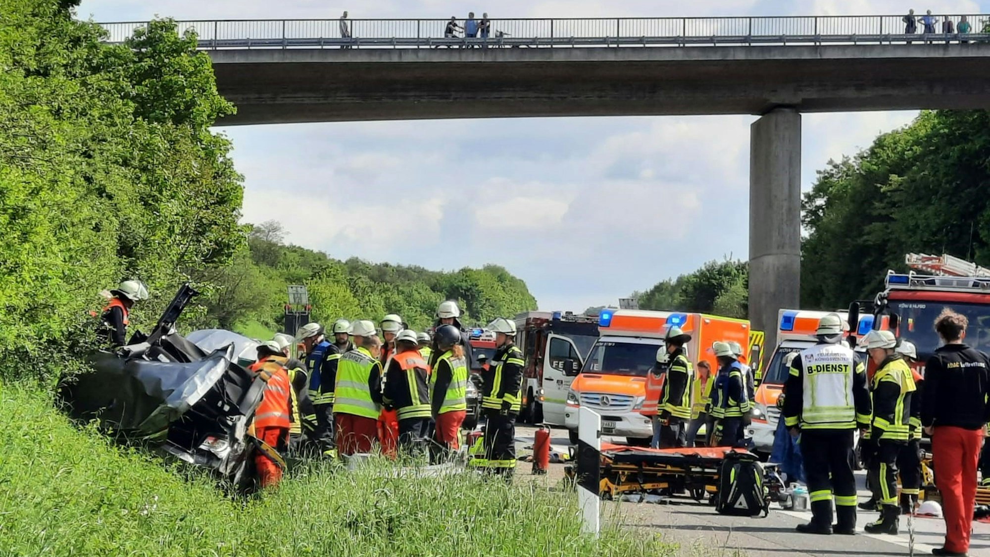 Dicht gedrängt stehen Rettungskräfte um ein Autowrack, das in der Böschung neben der Autobahn liegt. Auf einer Brücke stehen Schaulustige.