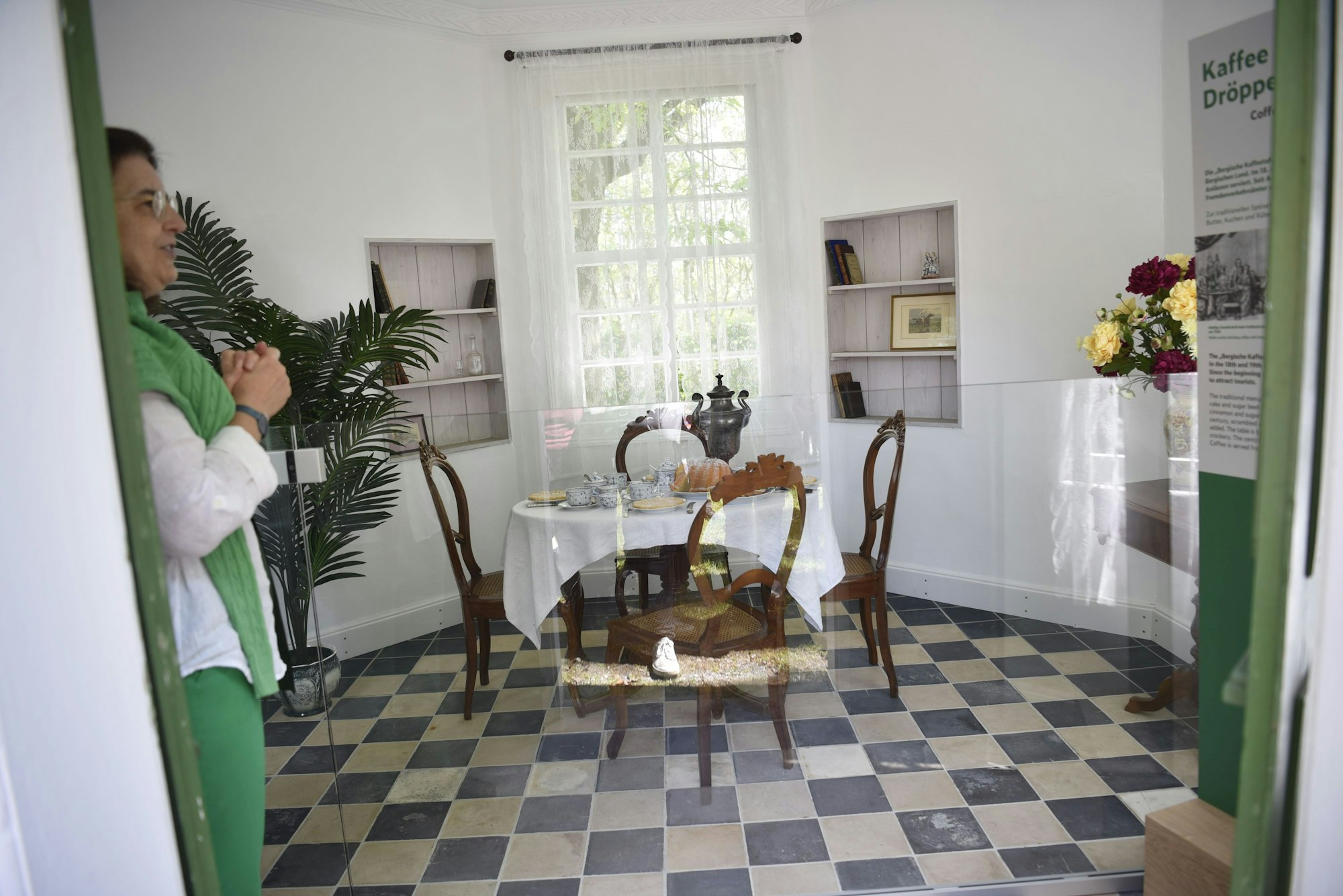 In einem fünfeckigen Raum stehen vor einem Fenster ein Tisch und vier Stühle. Auf dem Tisch liegt eine weiße Tischdecke. Der Tisch ist mit blau-weißem Geschirr gedeckt. Es sind ein Kuchen und eine Dröppelminna (eine alte Kaffeekanne) zu sehen. Am Rand stehen Blumen.