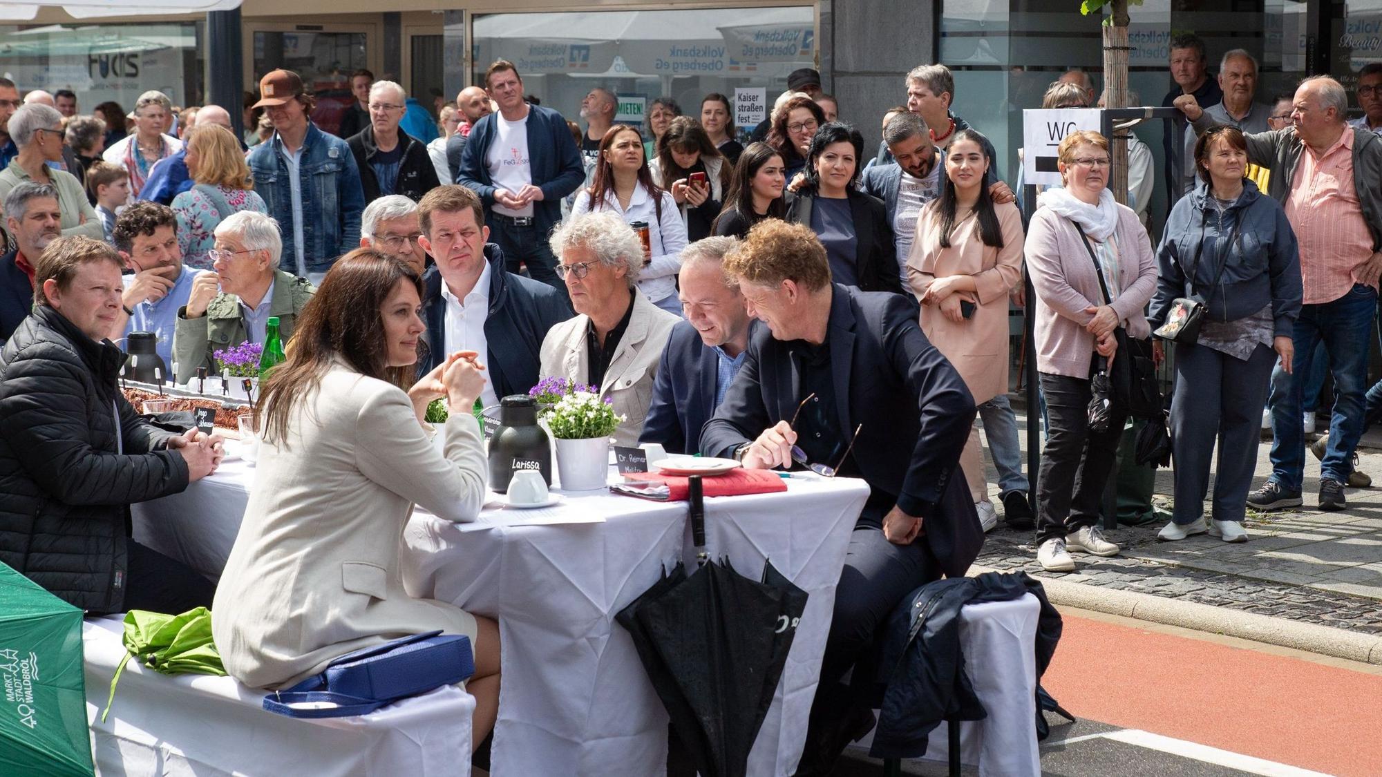 Zahlreiche Menschen säumen eine Straße in einem Stadtzentrum. In der Mitte steht eine lange weiße Tafel, an der ebenfalls viele Menschen sitzen.