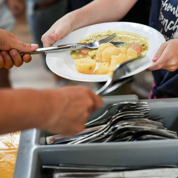 Das Foto zeigt Schüler beim Mittagessen in der Mensa in einer Grundschule. Im Vordergrund ist ein Besteckkasten zu sehen.