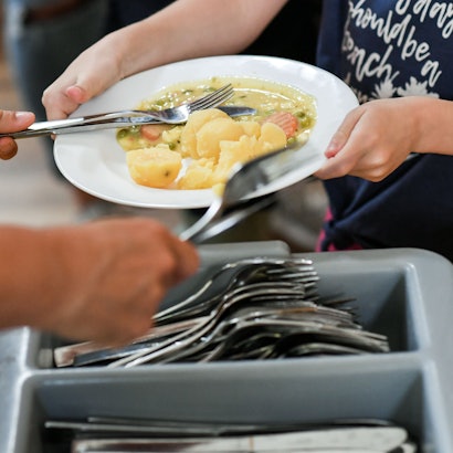 Das Foto zeigt Schüler beim Mittagessen in der Mensa in einer Grundschule. Im Vordergrund ist ein Besteckkasten zu sehen.