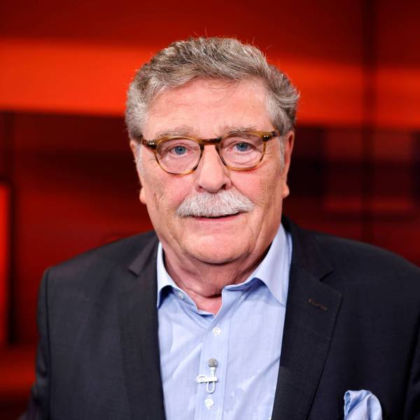 Der Politiker schaut in die Kamera, er trägt ein schwarzes Sakko und ein hellblaues Hemd. Der Hintergrund ist rot.