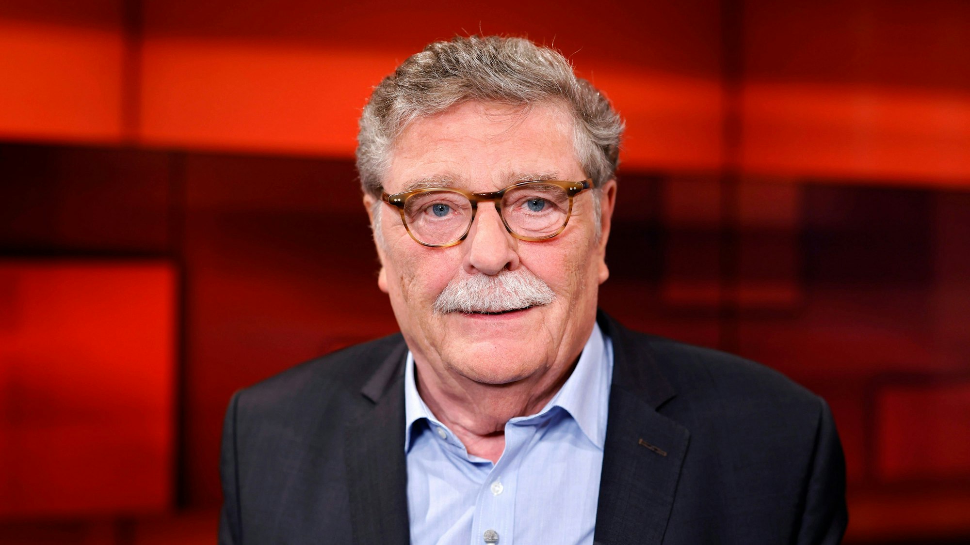Der Politiker schaut in die Kamera, er trägt ein schwarzes Sakko und ein hellblaues Hemd. Der Hintergrund ist rot.