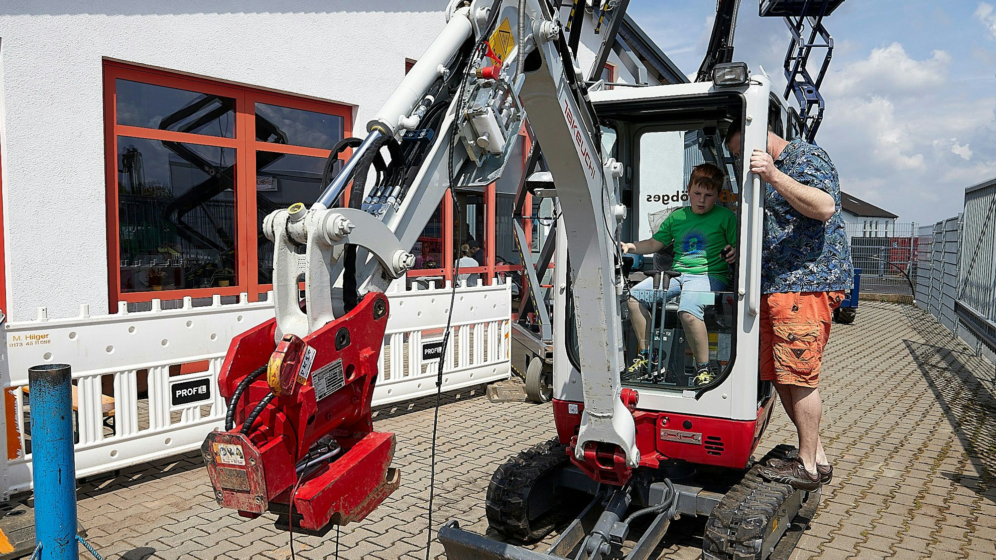 Der Junge bedient in der Kanzel die Steuerhebel des kleinen Baggers, während sein Vater in der Tür steht und Anweisungen gibt.