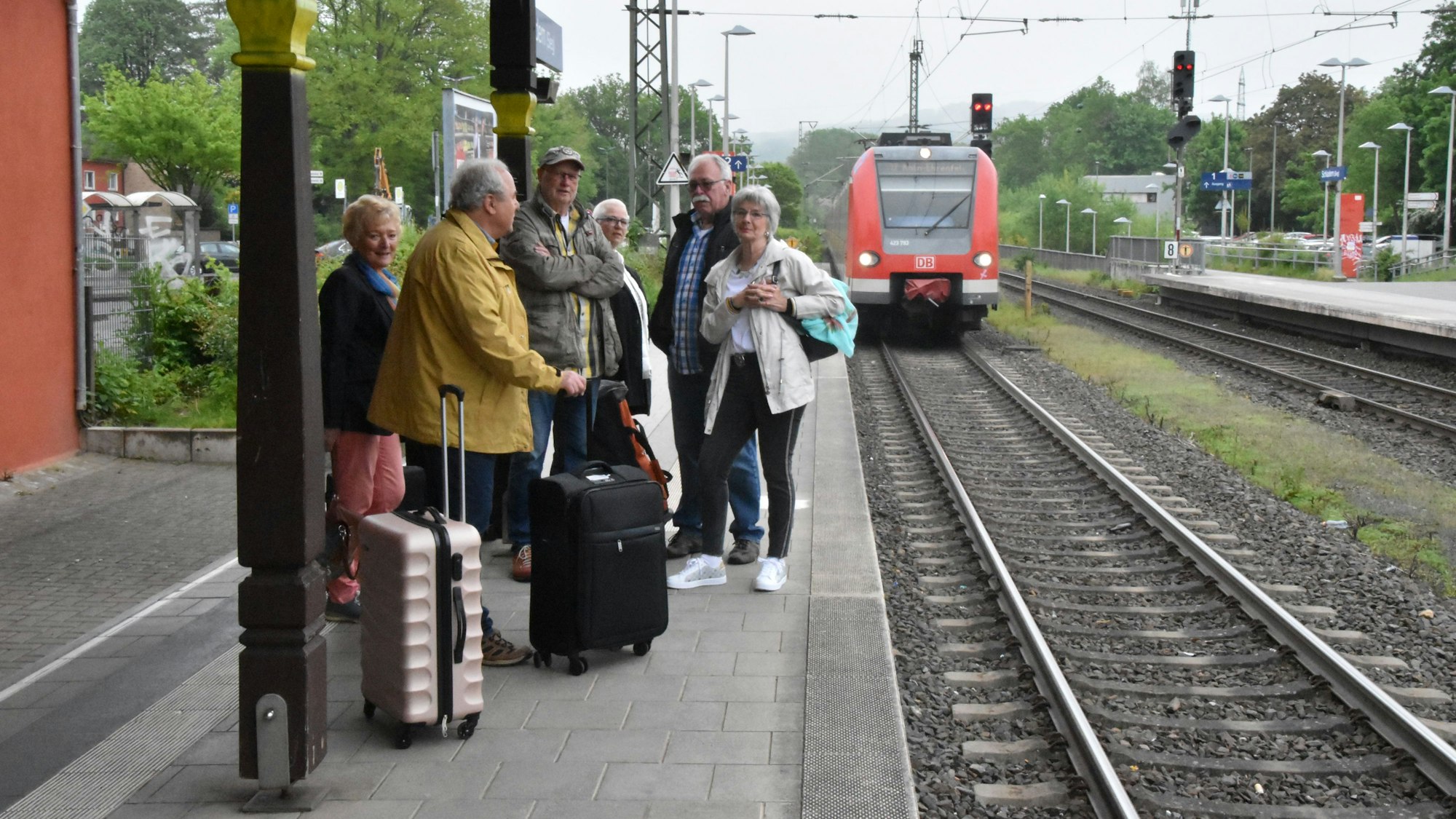 Auf einem Bahnsteig steht eine Gruppe Menschen mit Koffern, ein Zug fährt ein.