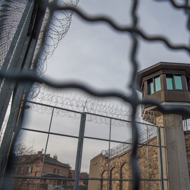 Mauern und Stacheldraht an einer JVA, im Hintergrund ist ein Wachturm zu sehen.