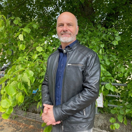 Marcus Venghaus trägt einen Ledermantel und steht vor einem grünen Baum.