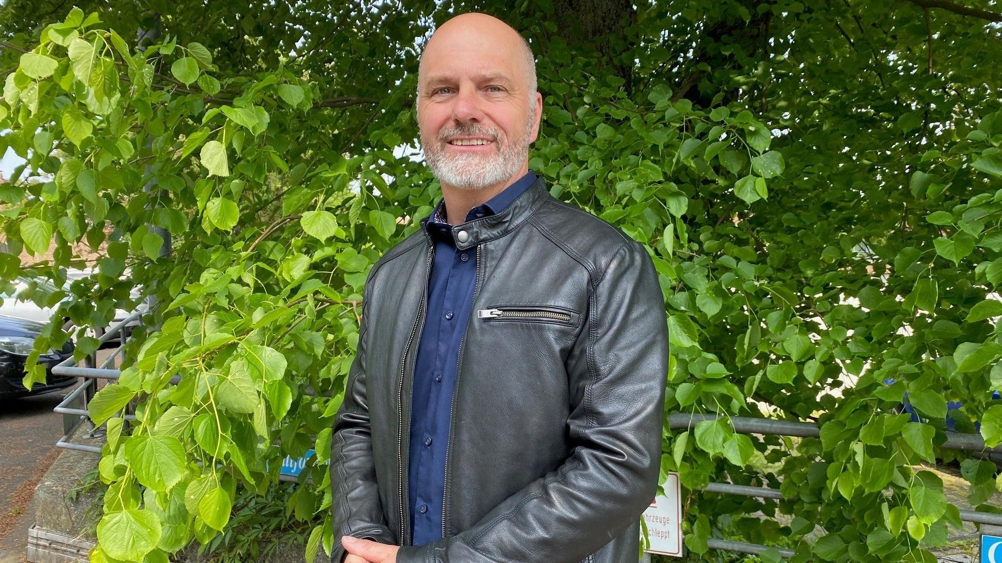 Marcus Venghaus trägt einen Ledermantel und steht vor einem grünen Baum.