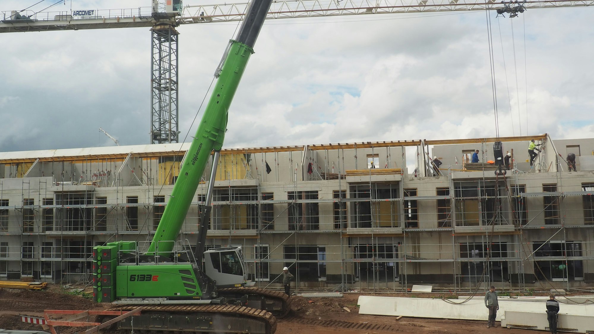Ein grünes Baustellenfahrzeug steht vor eine Reihe von Häusern im Rohbau. Dahinter ist ein Kran zu sehen.