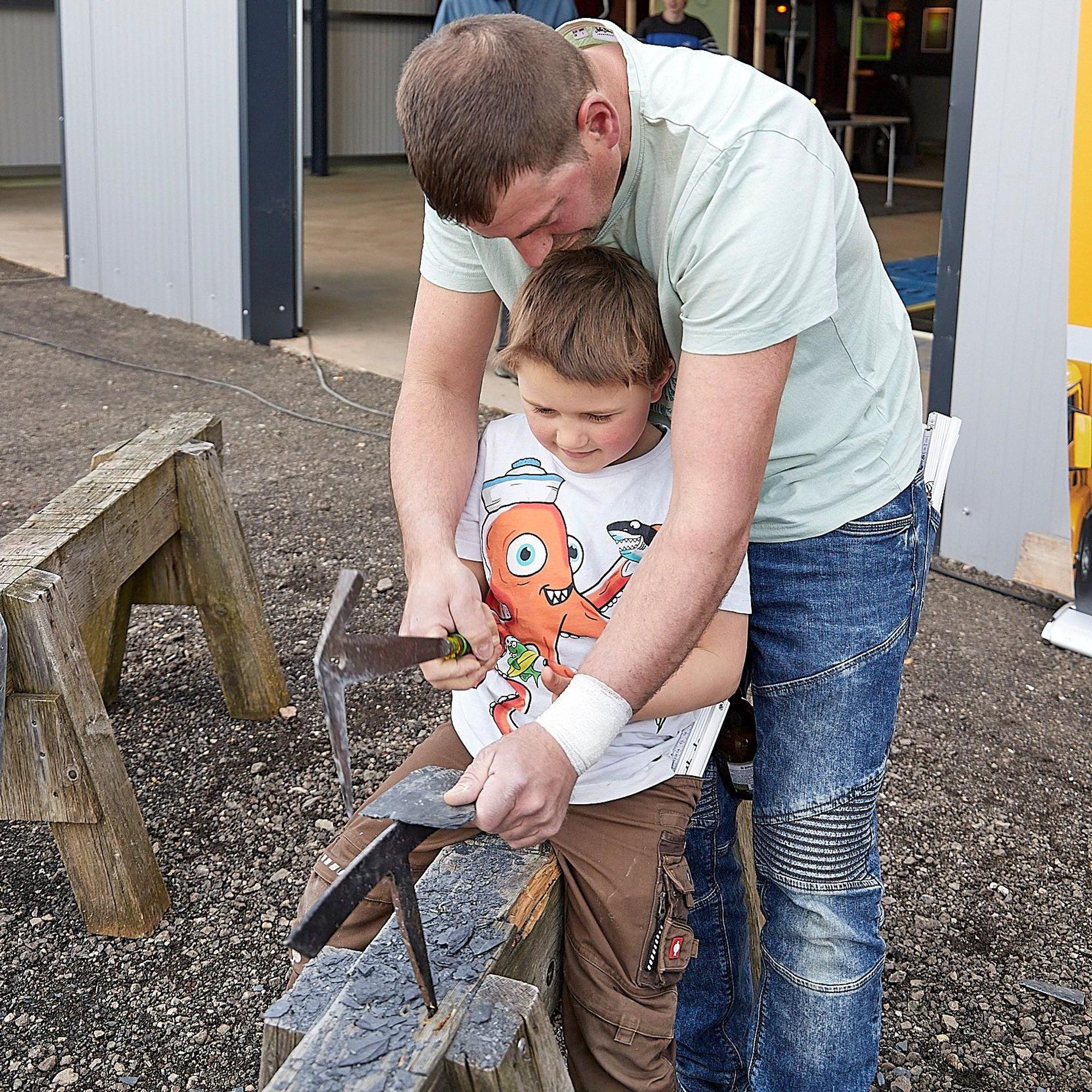 Der Vater zeigt dem Jungen, wie mit einem speziellen Hammer auf einem Holzbock der Schiefer in Form gebracht wird.