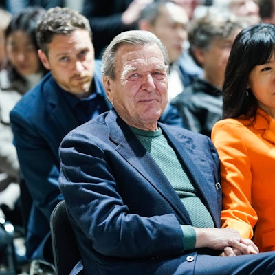 Der ehemalige Bundeskanzler Gerhard Schröder und seine Frau So-yeon Schröder-Kim sitzen im Publikum bei einer Veranstaltung. Frau Schröder-Kim trägt einen orangefarbenen Blazer.