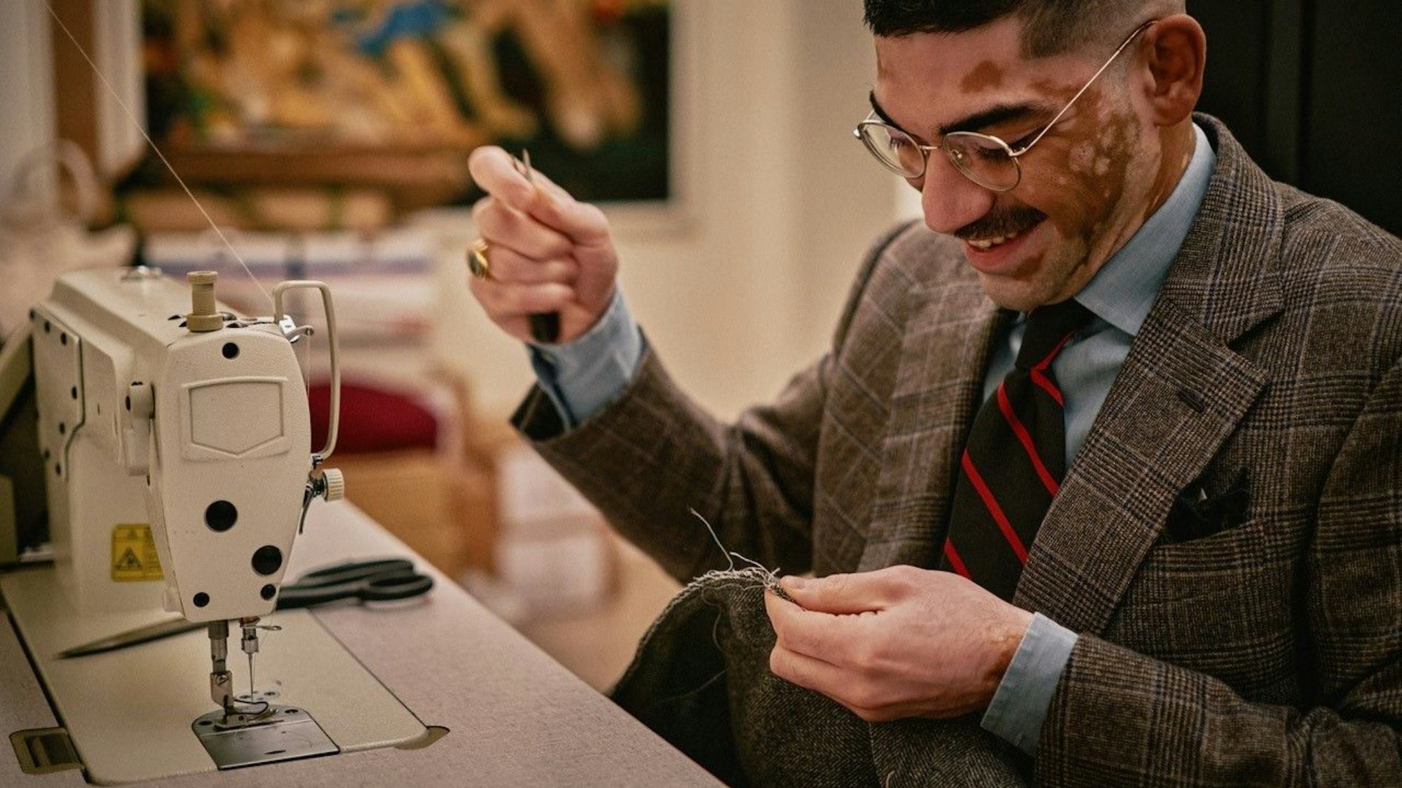 Ein Mann im Anzug sitzt vor einer Nähmaschine. Er hat Nadel und Faden in der einen und ein Stück Stoff in der anderen Hand.