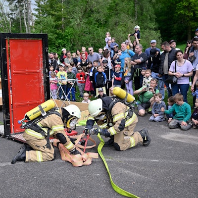 Feuerwehrleute montieren einen Feuerwehrschlauch. Im Hintergrund beobachten Zuschauer die Szenerie.