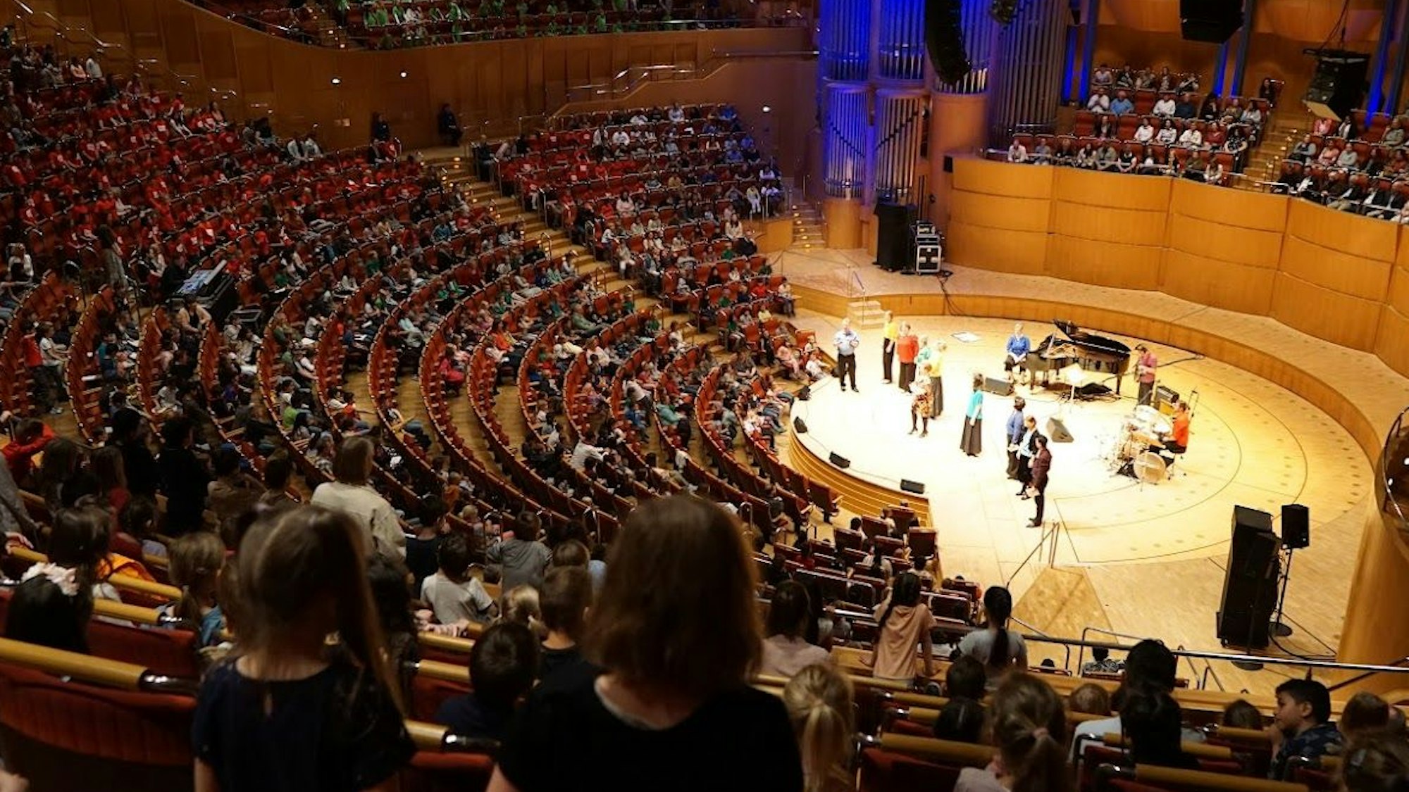 Die kleinen Sängerinnen und Sänger sitzen in den Zuschauerreihen, die Singleiter stehen auf der Bühne.