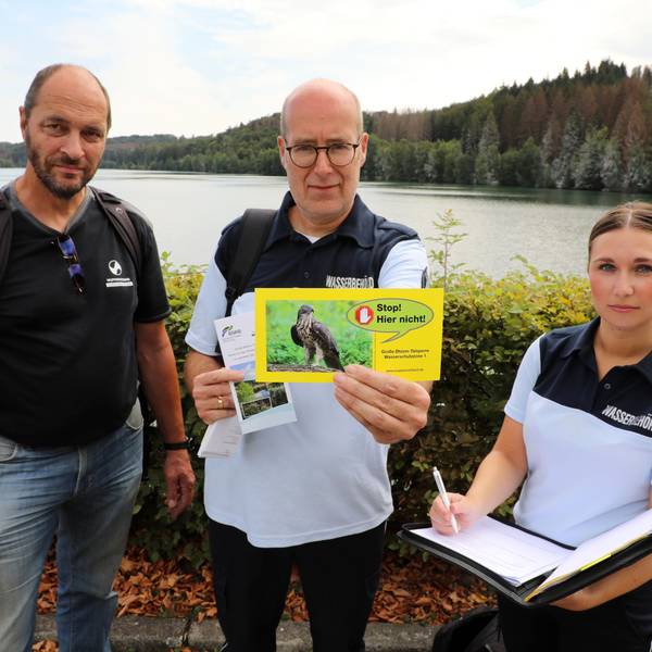 Zwei Männer und eine Frau stehen in Uniform vor einer Talsperre und halten eine gelbe Karte noch.