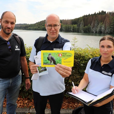 Zwei Männer und eine Frau stehen in Uniform vor einer Talsperre und halten eine gelbe Karte noch.