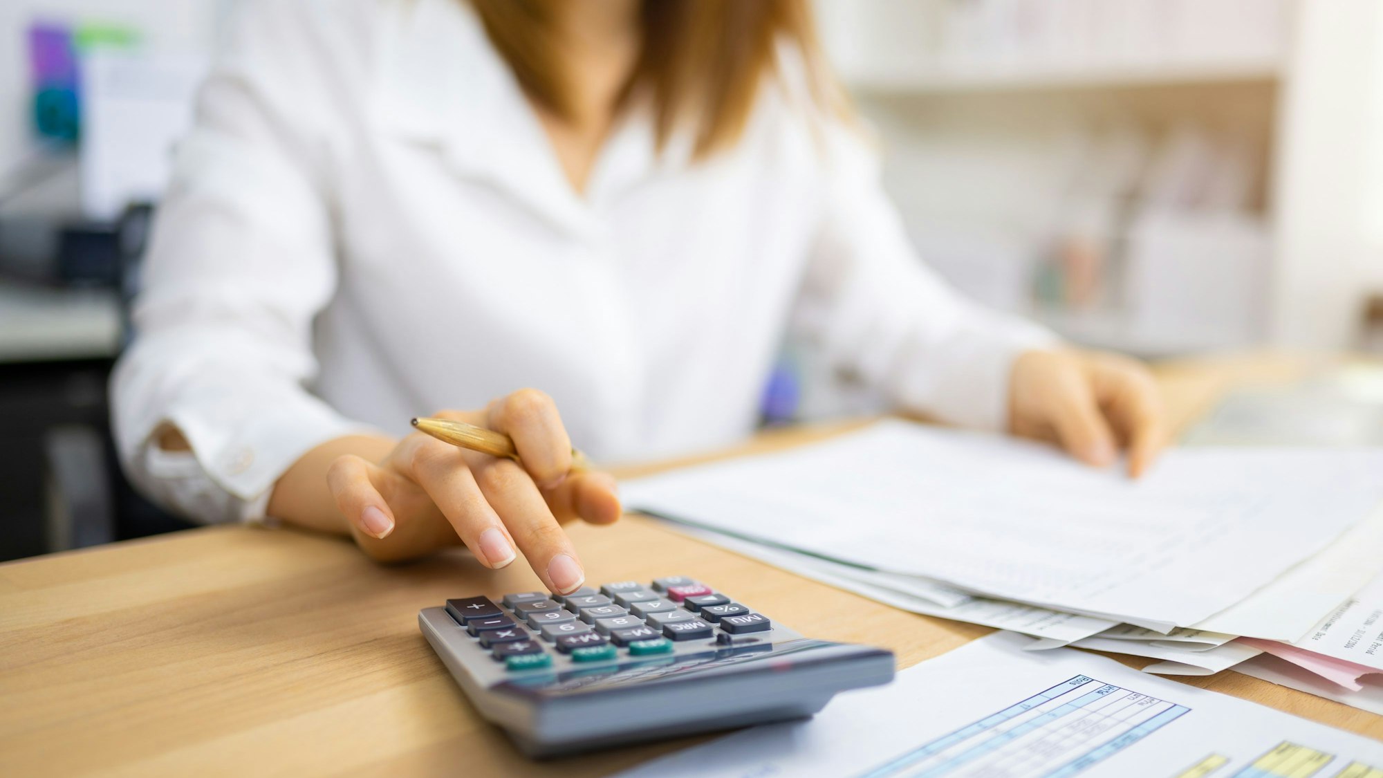 Unscharf zu sehen ist eine junge Frau am Schreibtisch, im Vordergrund ein Taschenrechner, Unterlagen und Formulare.