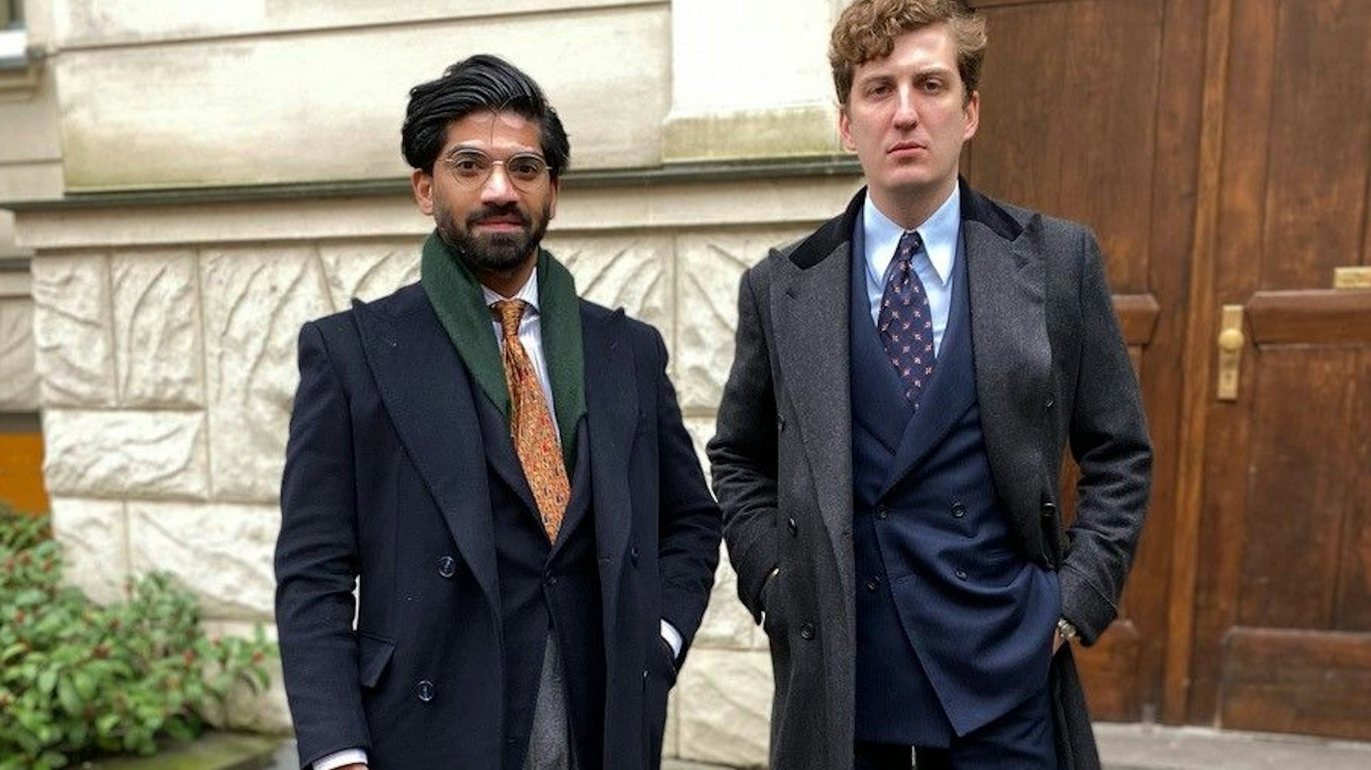 Zwei Männer in hochwertigen Mänteln stehen vor der Haustür eines Altbaus.