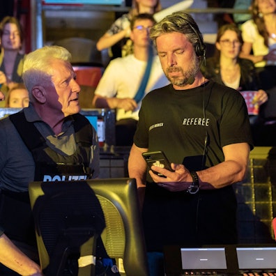 Die Ermittler haben schusssichere Westen mit der Aufschrift „Polizei“ an, der Schiedsrichter ein T-Shirt mit der Aufschrift: „Referee“. Er schaut auf ein Handy, dass die Ermittler ihm überreicht haben. Im Hintergrund Zuschauer in einer Gaming-Arena.