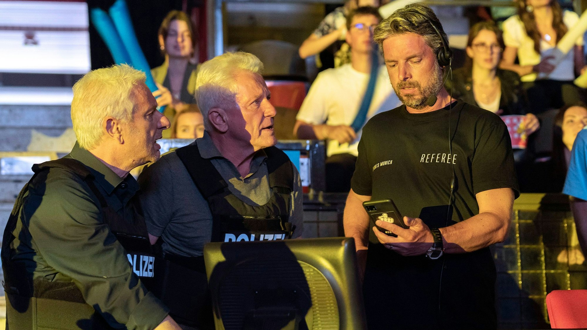 Die Ermittler haben schusssichere Westen mit der Aufschrift „Polizei“ an, der Schiedsrichter ein T-Shirt mit der Aufschrift: „Referee“. Er schaut auf ein Handy, dass die Ermittler ihm überreicht haben. Im Hintergrund Zuschauer in einer Gaming-Arena.