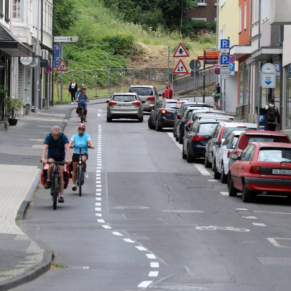 Links auf dem Foto sind Radfahrer auf dem Radstreifen unterwegs. Auf dem Parkstreifen rechts im Bild parken Autos. Auf der Fahrbahn in der Mitte fahren Autos.