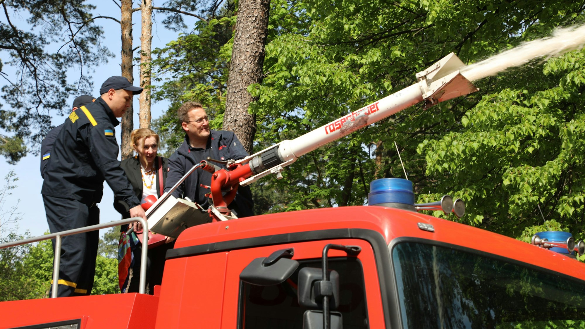 Drei Feuerwehrleute und eine Dolmetscherin stehen auf einem Tanklöschfahrzeug und bedienen den Wasserwerfer auf dem Dach des Feuerwehrautos.