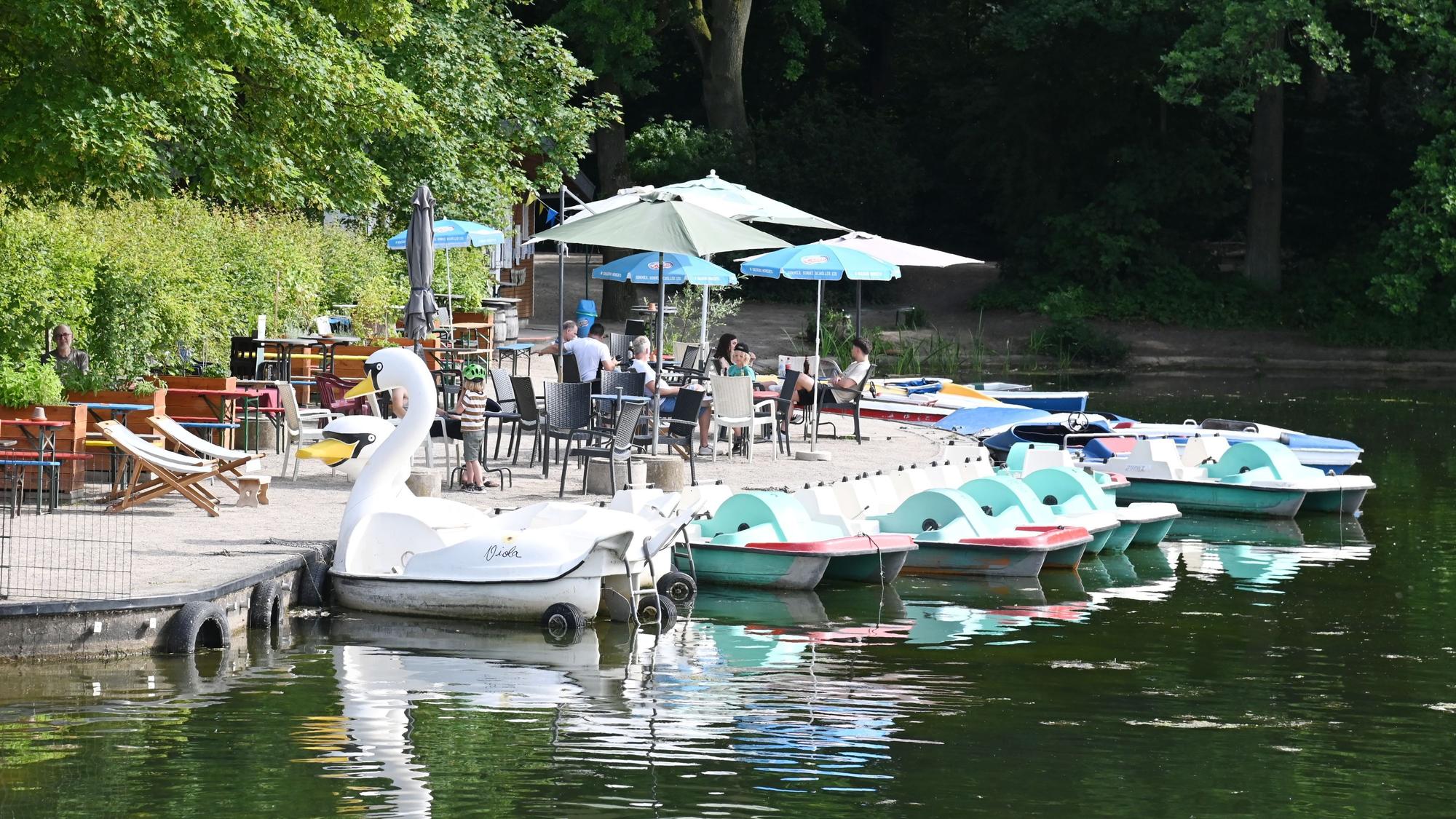 Terrasse mit Sonnenschirmen und Liegestühlen am Rand eines Weihers, auf dem Wasser liegen mehrere Tretboote, unter anderem in Form eines Schwans