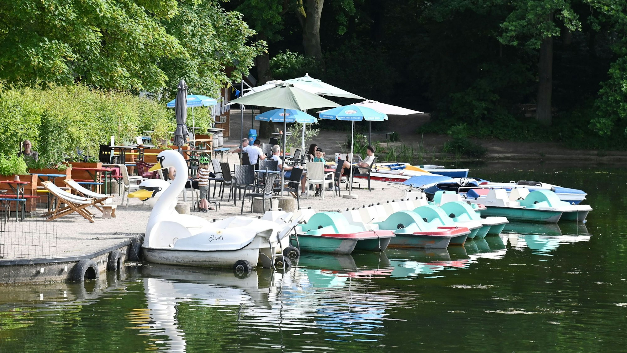 Terrasse mit Sonnenschirmen und Liegestühlen am Rand eines Weihers, auf dem Wasser liegen mehrere Tretboote, unter anderem in Form eines Schwans