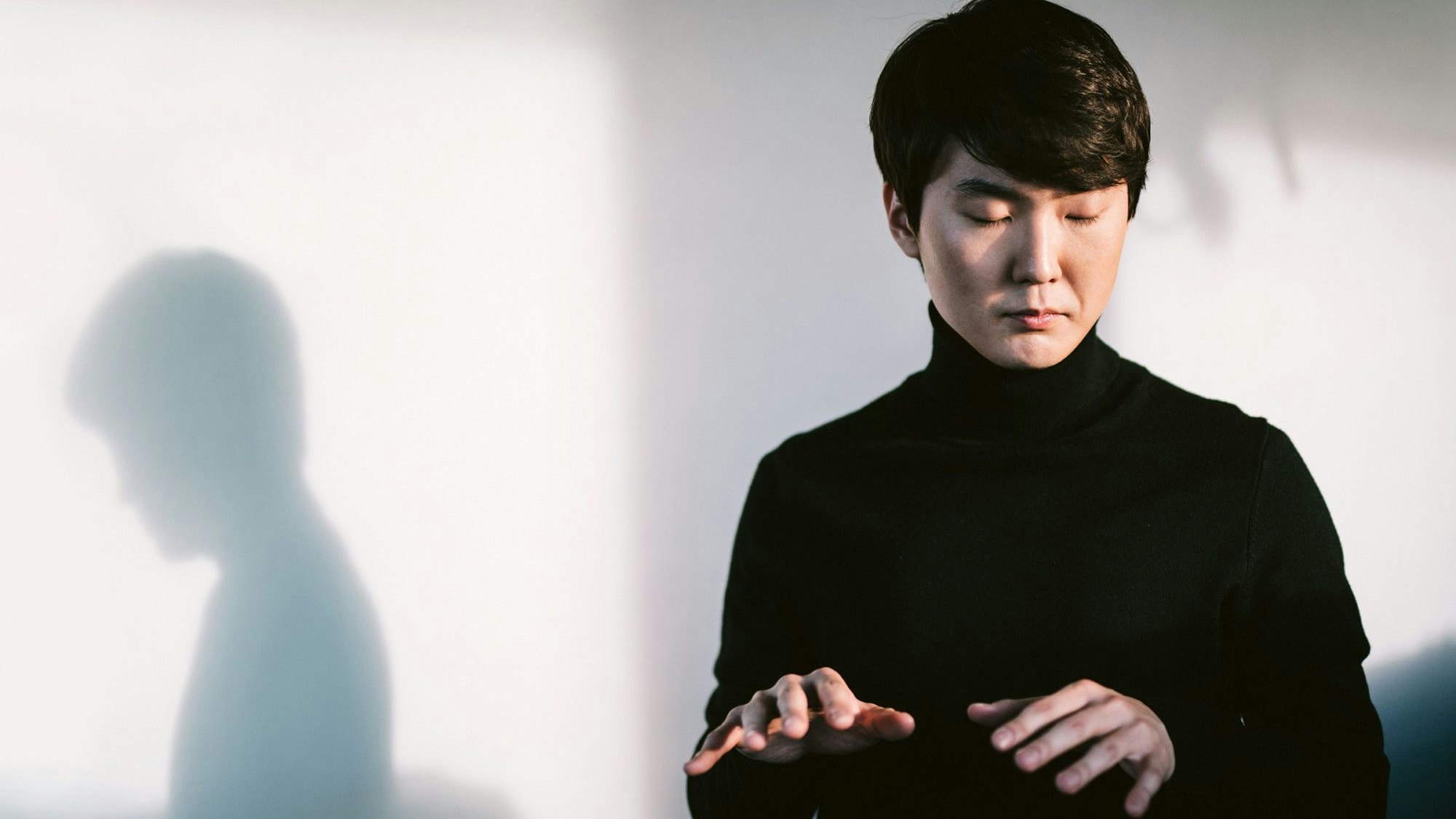 Pianist Seong-Jin Cho trägt einen schwarzen Rollkragen-Pullover und hält die Hände in die Luft, als würde er Klavier spielen.