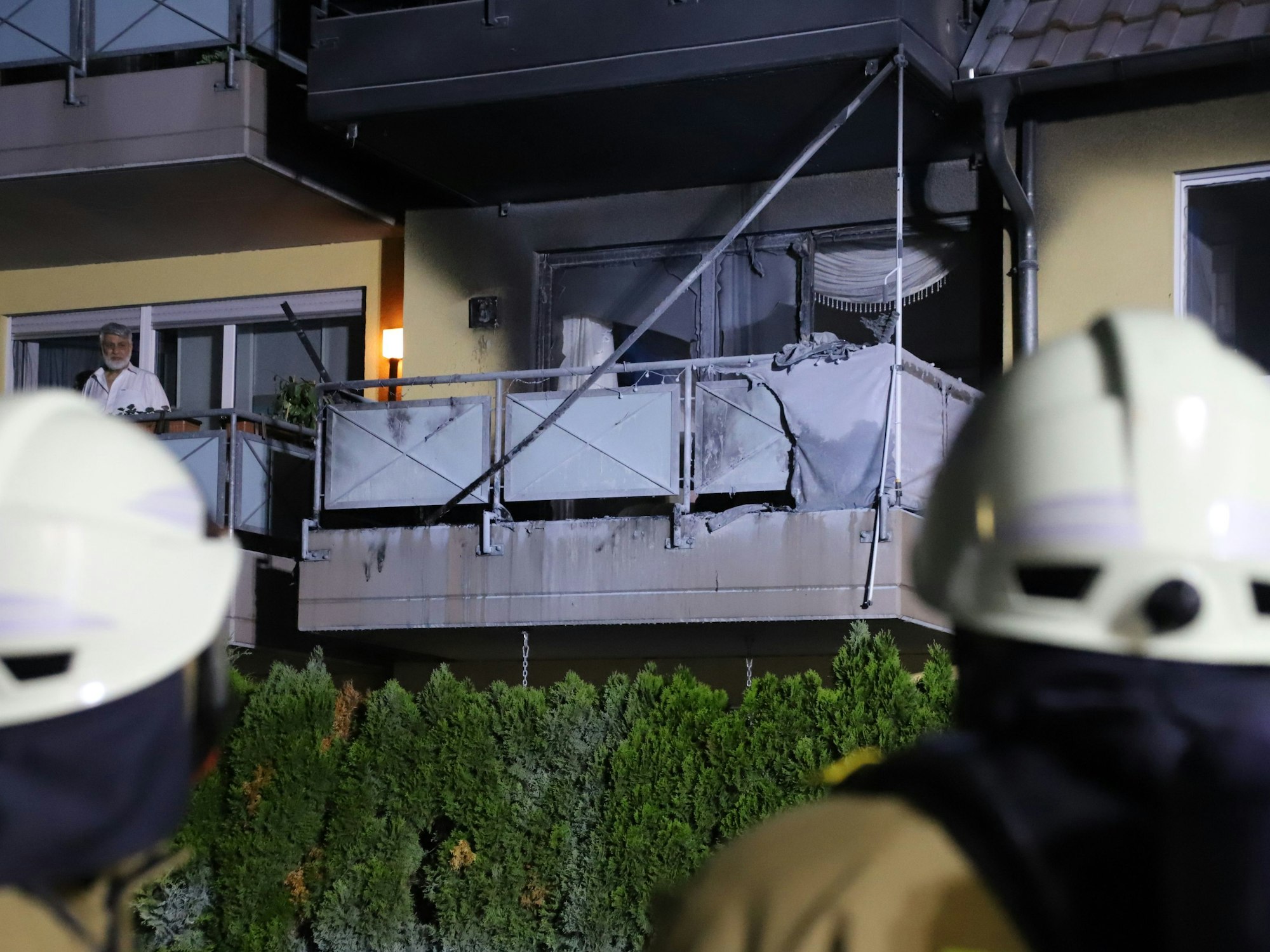 Ein Mann steht auf dem Balkon neben dem verbrannten Balkon. Im Vordergrund Feuerwehrleute.