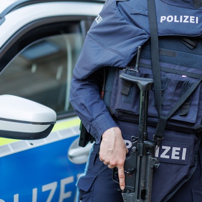 Eine Polizistin steht mit ballistischem Schutzhelm, einer Schusswaffe und einer kugelsicheren Weste vor einem Polizeiwagen.