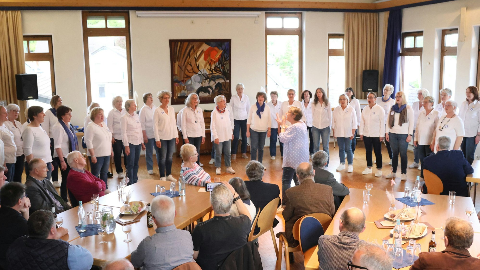 Zu sehen ist ein Frauenchor, der vor sitzenden Gästen singt.