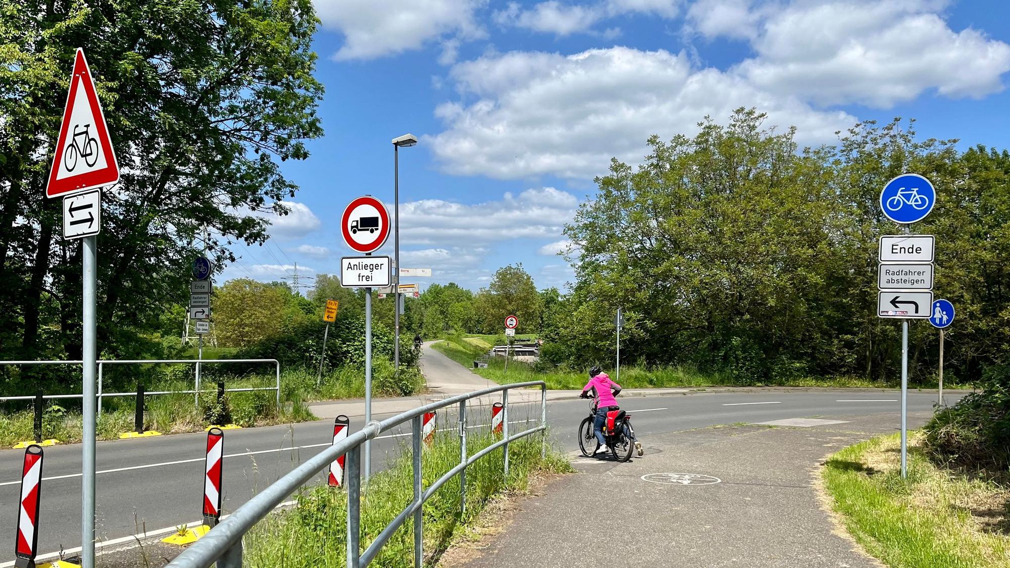 An der Einmündung der Rheindorfer Straße in den Westring widersprechen sich die Verkehrsschilder. Eine Radlerin wagt es, nicht abzusteigen.
