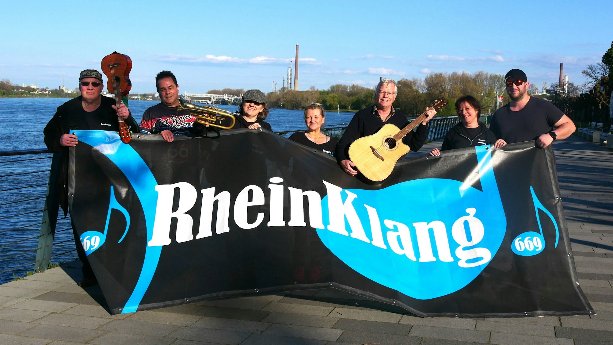 Das Organisationsteam des Festivals RheinKlang 669 posiert mit Instrumenten und dem Banner „RheinKlang“.