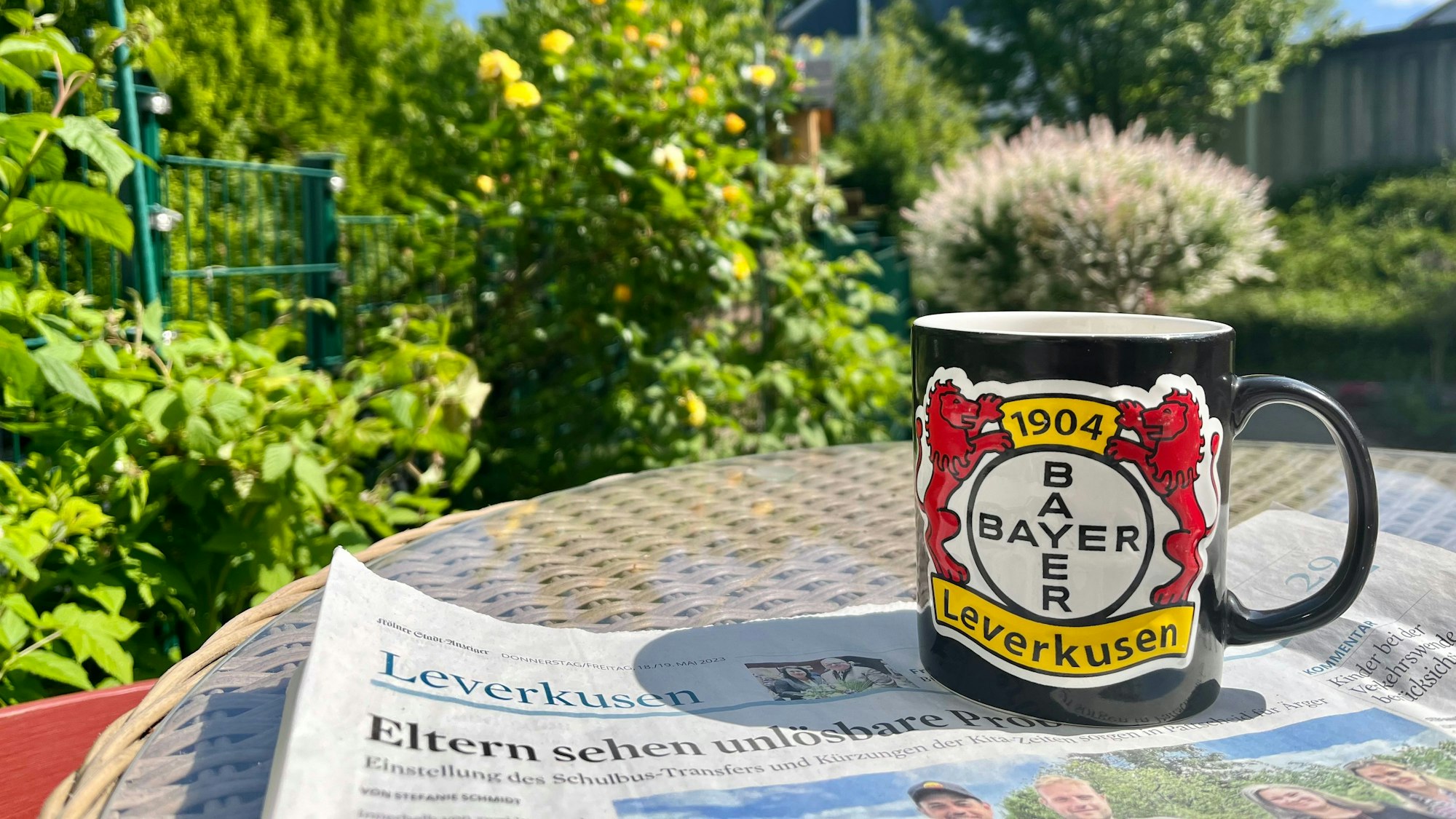 Auf einem Gartentisch liegt eine Ausgabe des „Leverkusener Anzeiger“, darauf stehe eine Bayer-04-Tasse.
